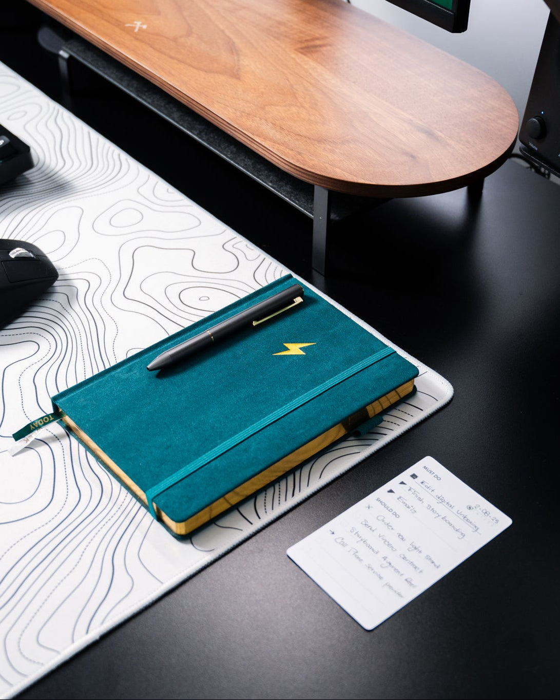 Teal notebook with a pen on a desk with a keyboard, mouse, and monitor.
