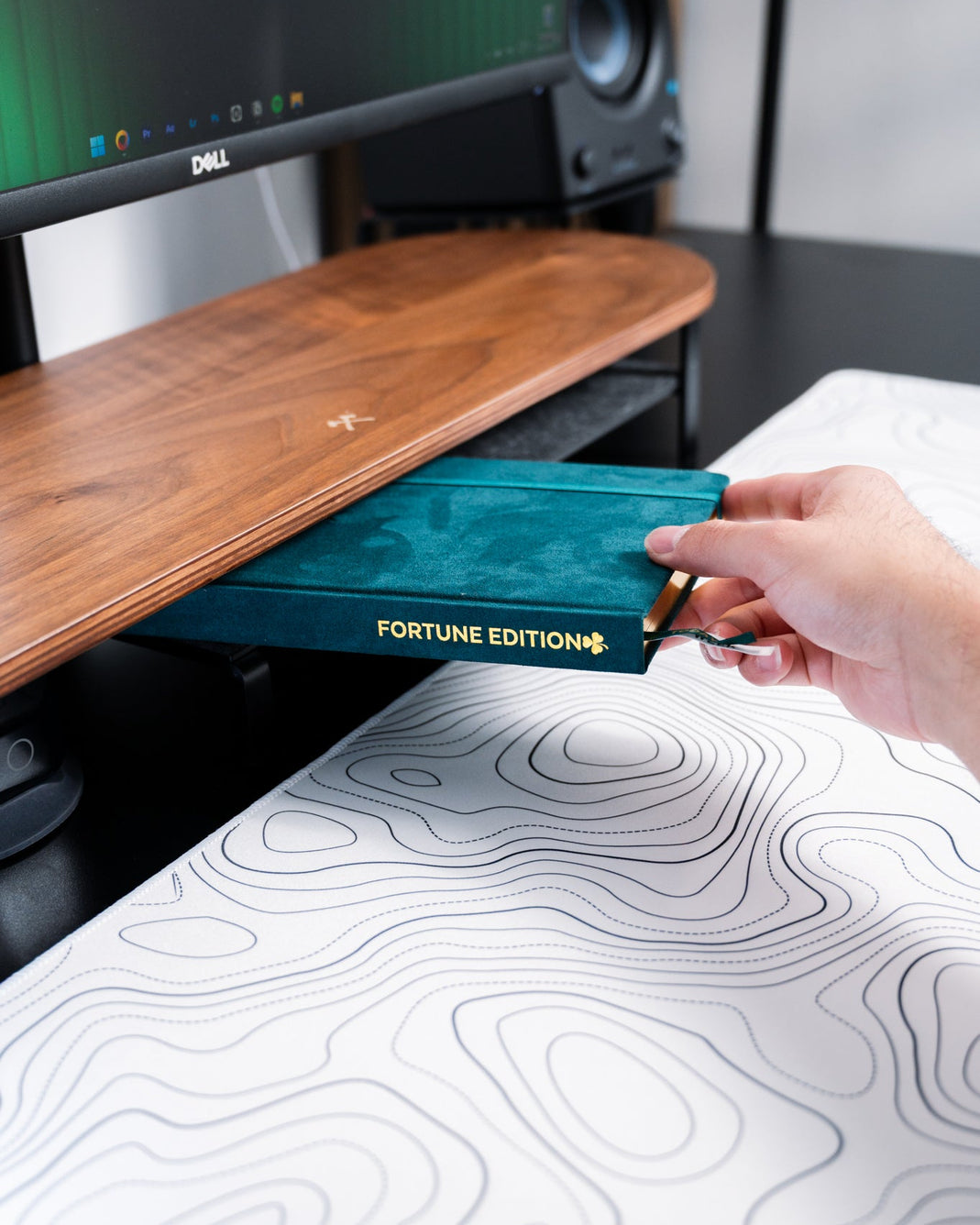 Hand holding a green book titled 'Fortune Edition' in front of computer monitors.
