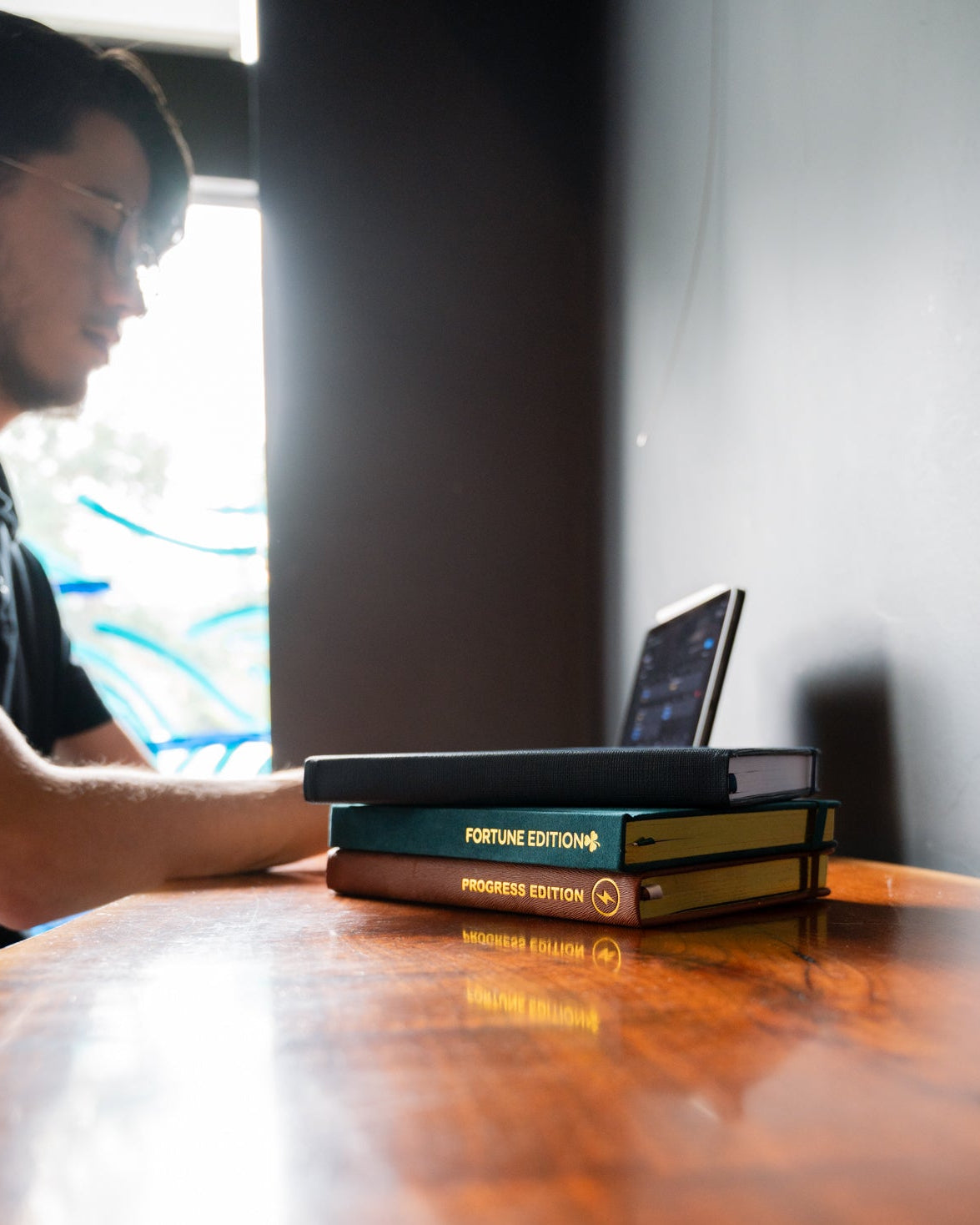 Stack of books on a wooden table with a person sitting in the background