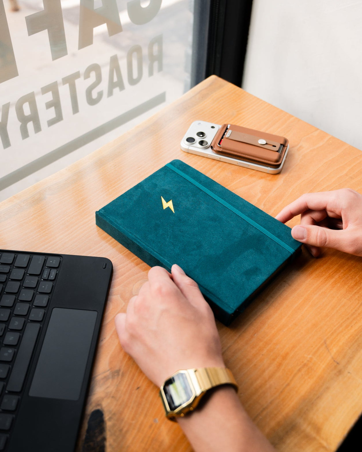 Teal wallet with gold emblem on a wooden table next to a phone and keyboard, with a blurred background.