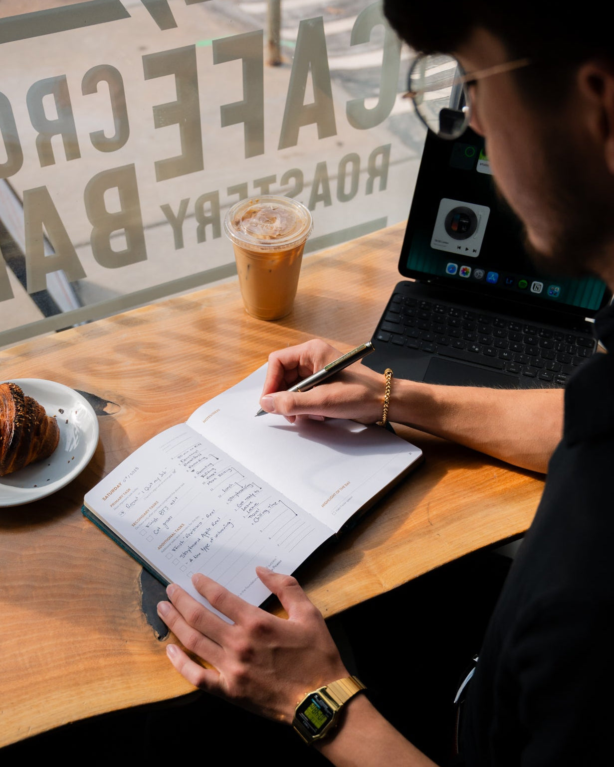 Person sitting at a table with a laptop and a drink, in front of a coffee shop window.